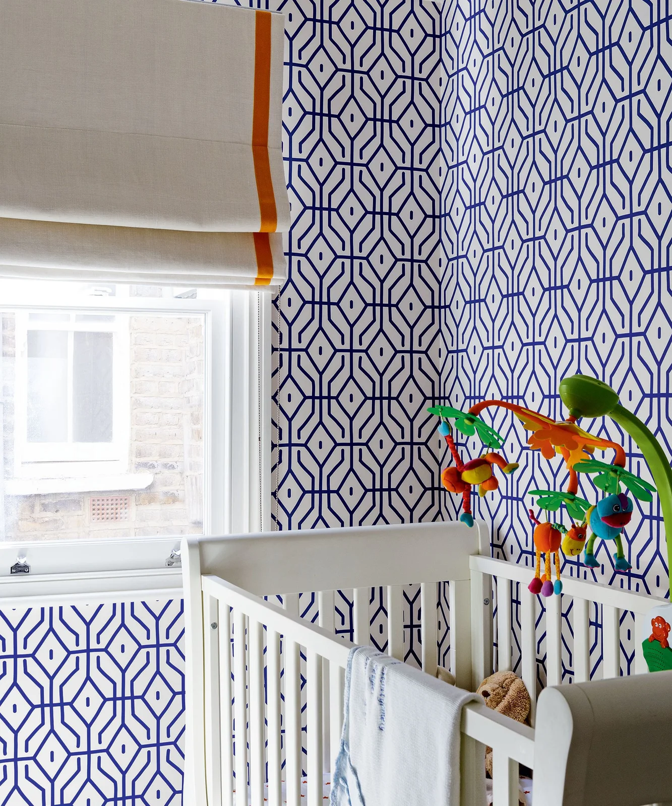 Blue geometric wallpaper surrounds a white nursery crib beside a pale blind with an orange border and colorful hanging mobile