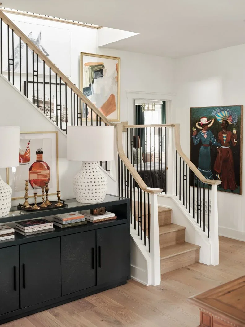 A dark sideboard with lower doors and an open upper shelf stands beneath stairs beside two pale lamps, framed artwork and stacked books