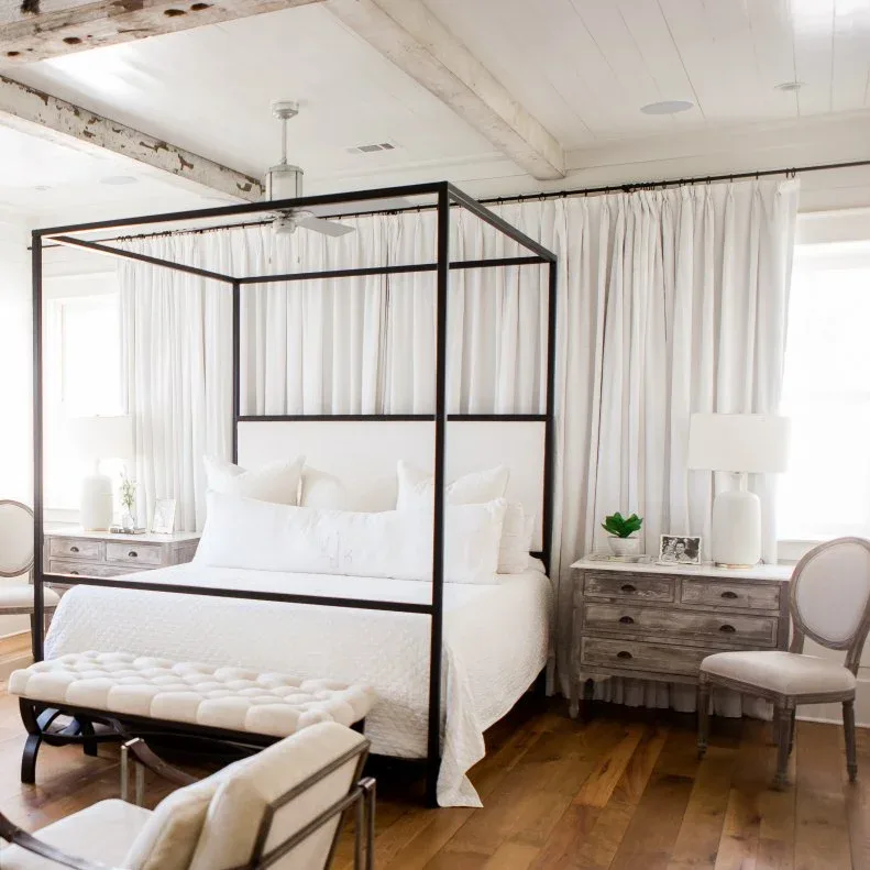 White bedroom with dark canopy bed, pale curtains, weathered bedside drawers and upholstered bench