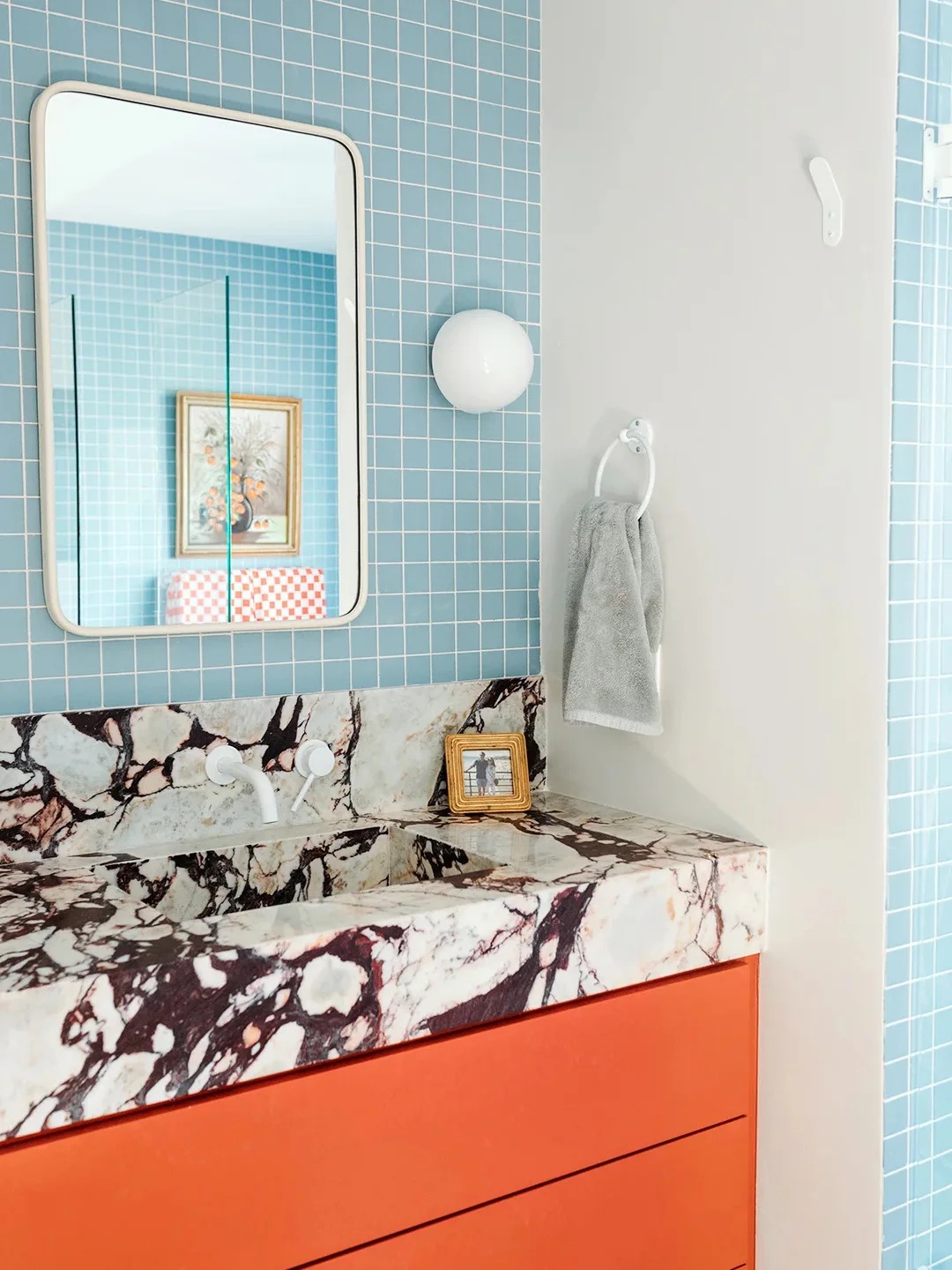 An orange bathroom vanity stands beneath a boldly veined counter, a rounded rectangular mirror and white globe light beside pale blue square tiles