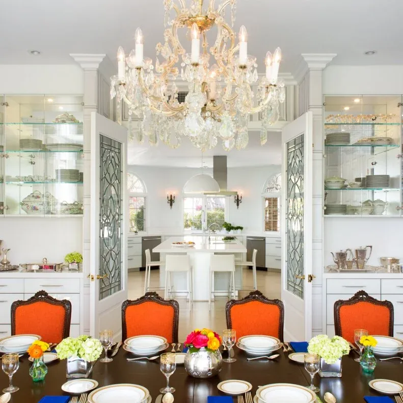 White glazed display cabinets on both sides of open decorative doors, with lower drawers, a dining table, orange upholstered chairs, and ornate lighting.