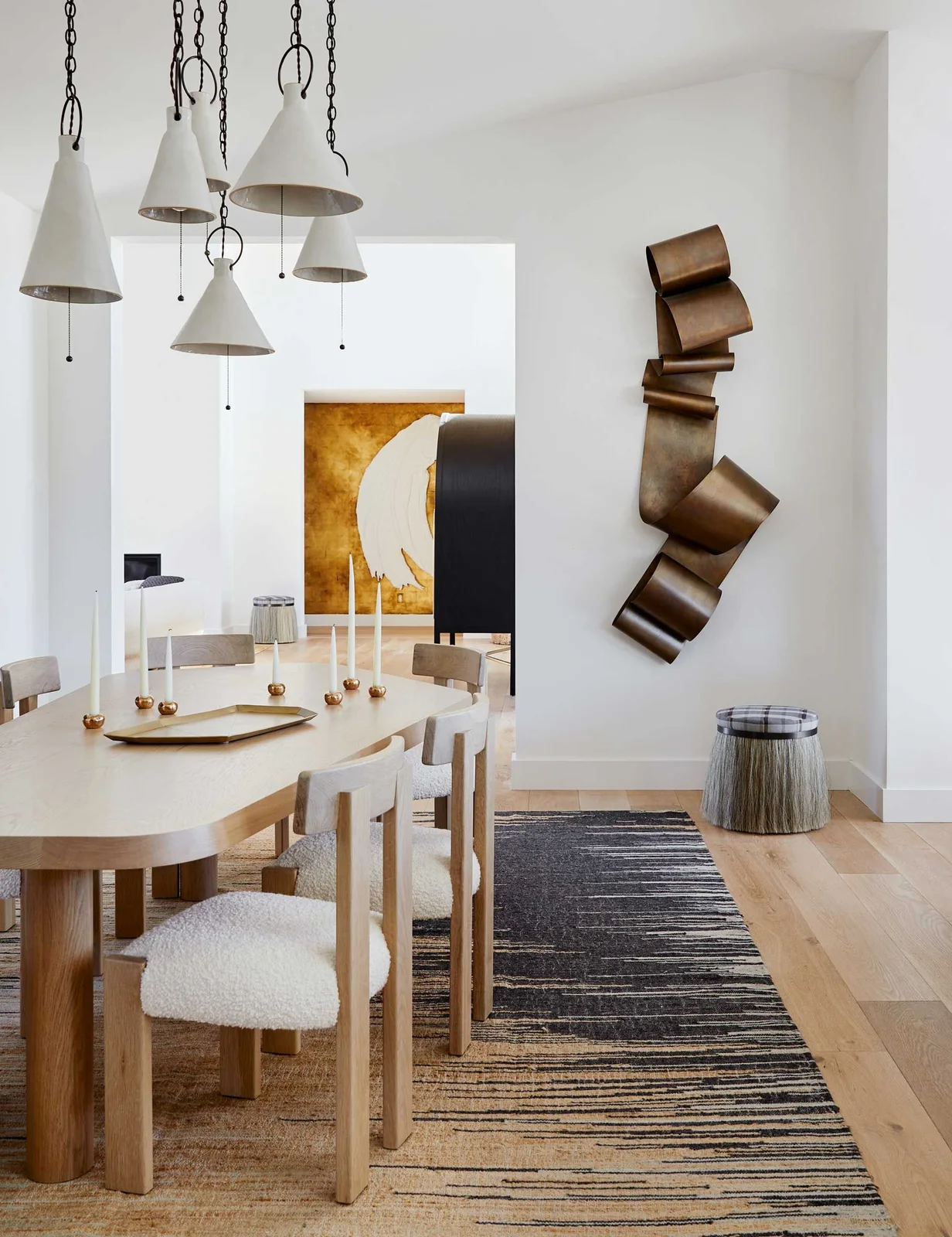 A pale dining table with an irregular outline and broad wood supports stands among pale upholstered chairs beneath white cone lights beside abstract art and a striped rug