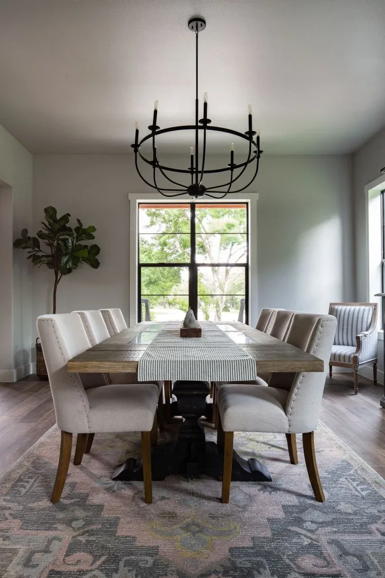 Black open circular chandelier above a long wood table with pale upholstered chairs and a striped runner