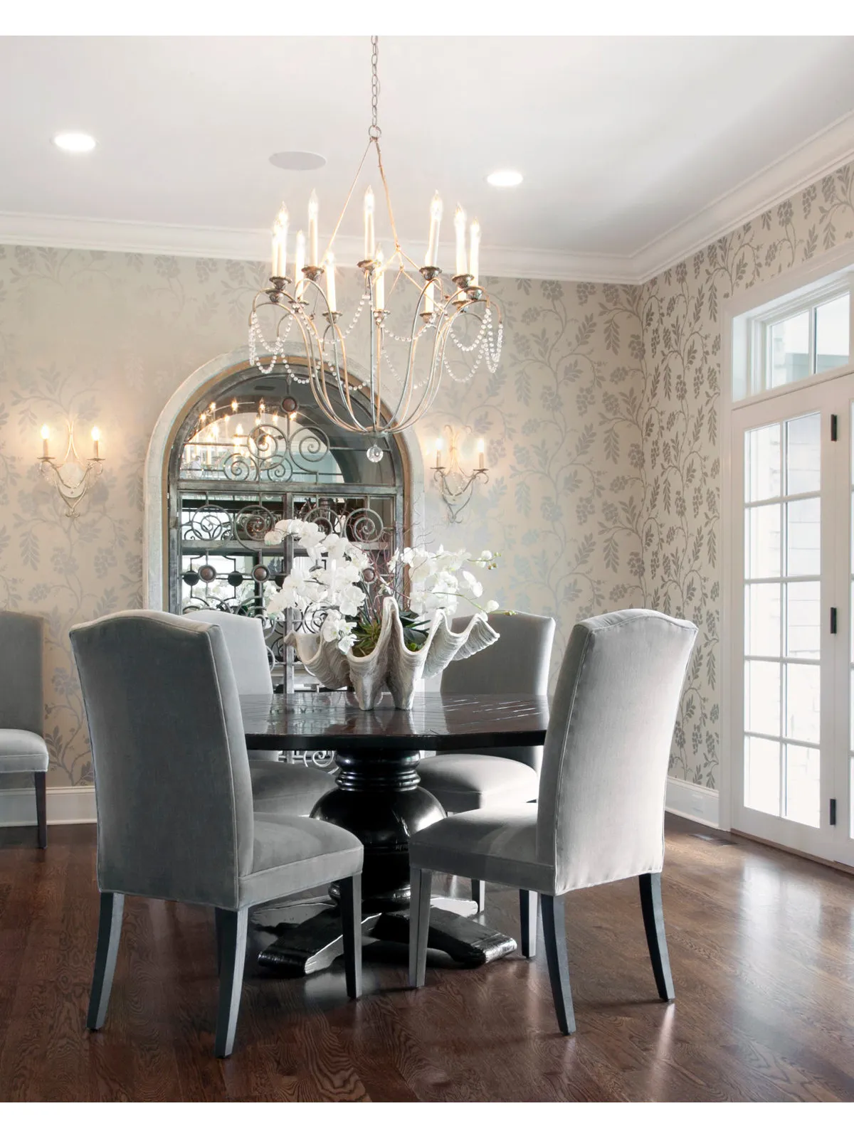 Tall arched mirror against softly patterned walls behind a dark table and pale upholstered chairs