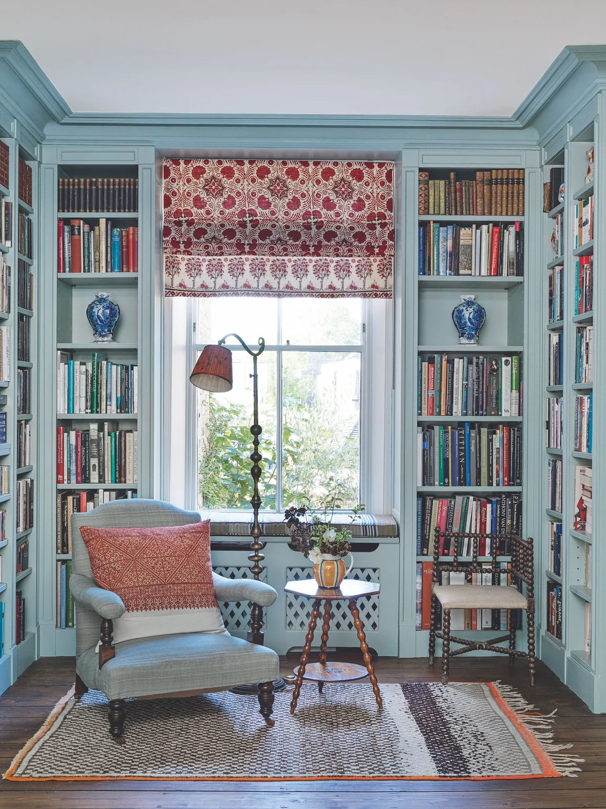 Pale blue bookcases surround a window with a red patterned blind beside a blue armchair, floor lamp and small wooden side table