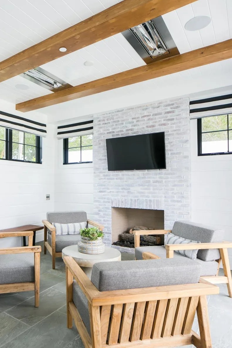Pale brick fireplace under a television with wooden-framed gray chairs, striped blinds and ceiling beams.