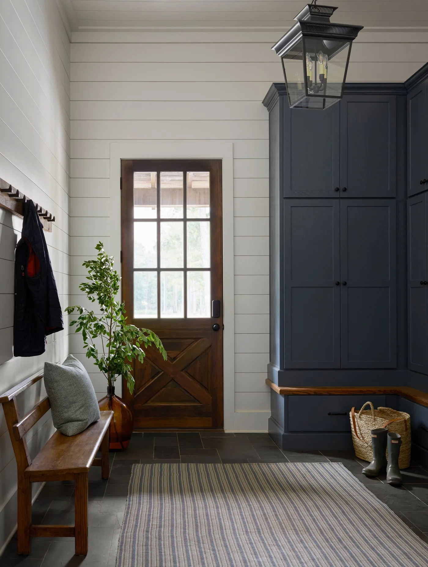 Black glazed candle lantern in white horizontal-paneled entry with wood door, dark blue cupboards, wood bench and striped runner
