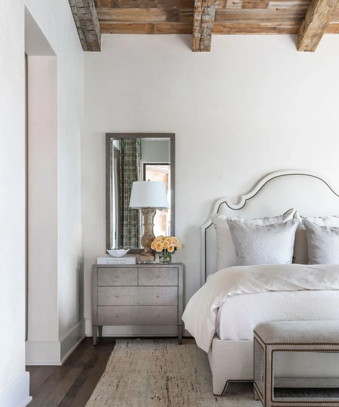 Pale bedroom with exposed wooden ceiling beams, white shaped headboard, gray bedside drawers, tall rectangular mirror and a light bench.