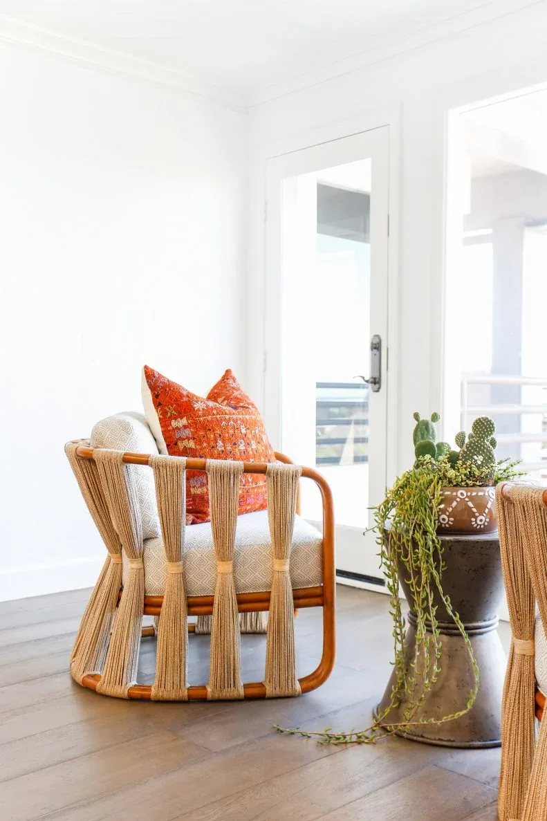 Pale living room corner with sculptural woven chair, light seat cushion, orange accent pillow and several potted plants beside a glazed door.