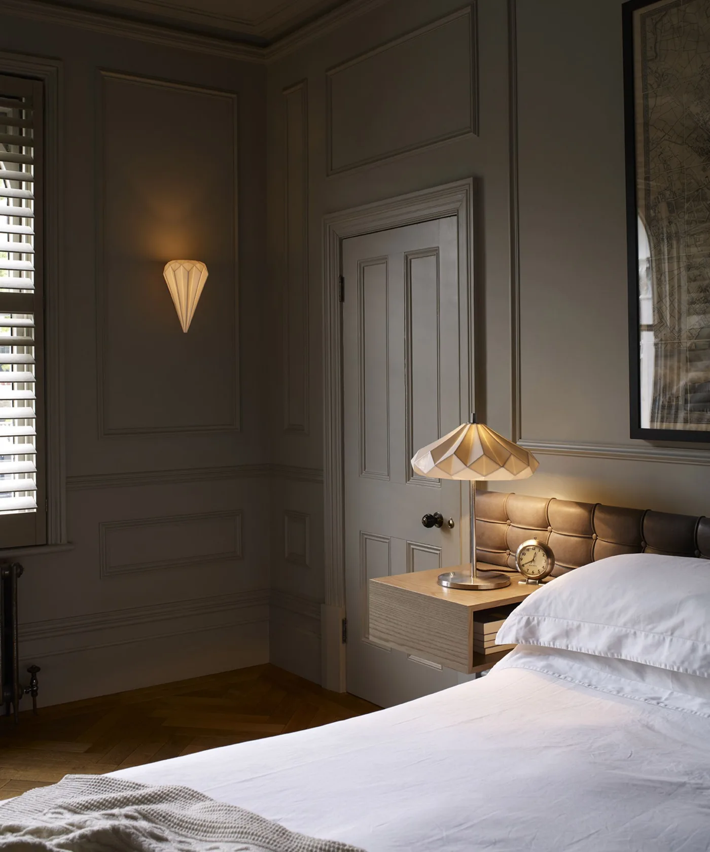 Bedroom with dark gray wall paneling, pale bedding, wooden bedside cabinet, warm table lamp and small wall light beside the door.