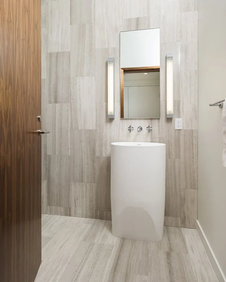 A tall white freestanding basin stands beneath a narrow rectangular mirror between slim wall lights against vertically patterned pale gray bathroom tiles