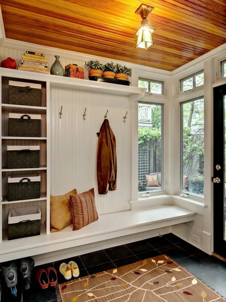 White board-lined mudroom with long white bench, hooks, baskets on shelves, dark floor, wood ceiling and corner windows