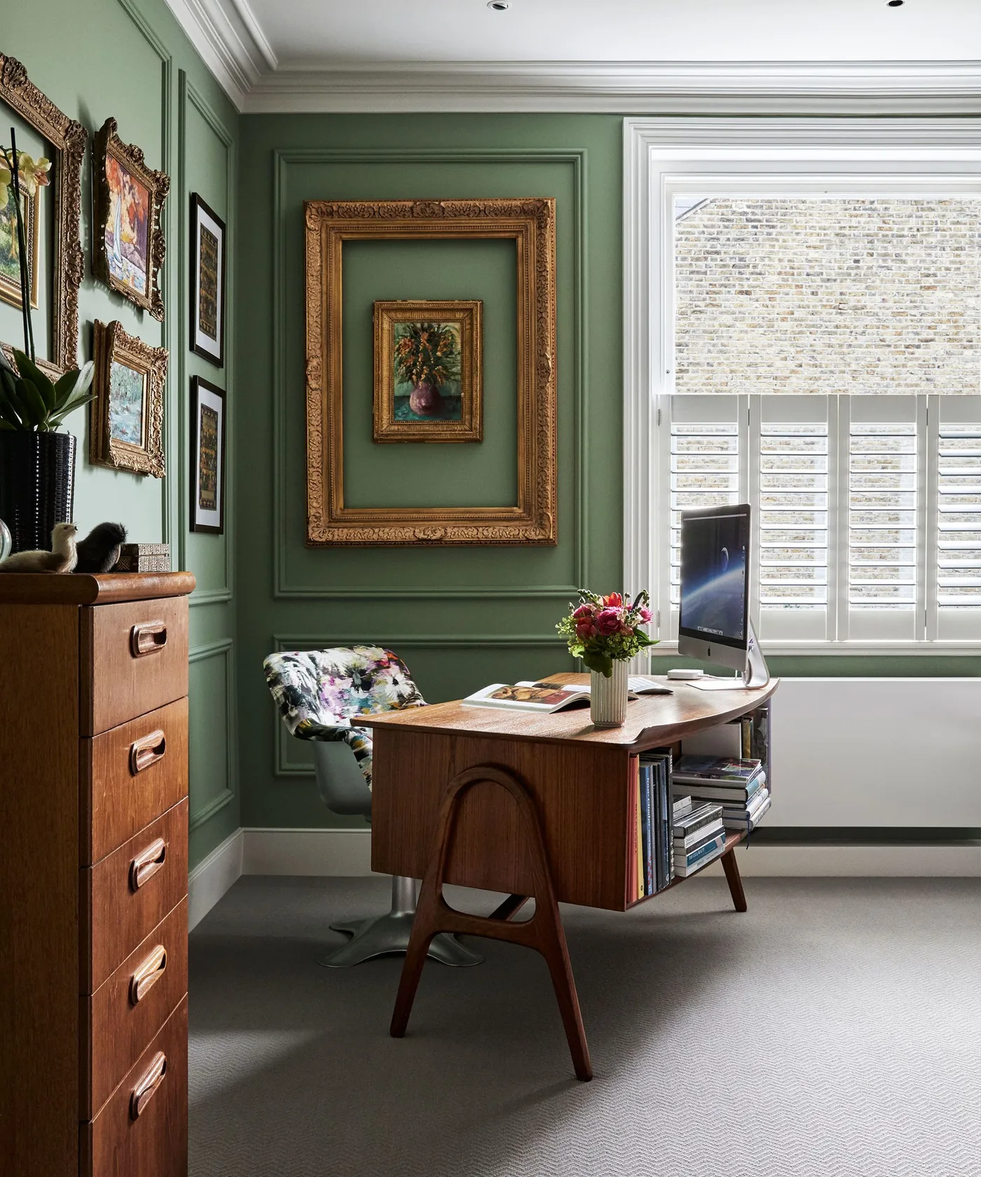 Green paneled office with ornate gold-toned picture frames, a wooden desk and a bright window.