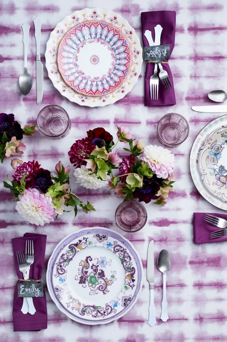 Purple napkins and pale purple glasses sit beside patterned plates with small dark name cards and light-colored cutlery