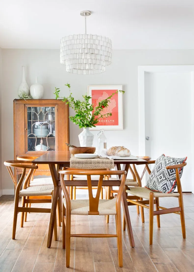 Pale tiered chandelier made of repeated rectangular pieces above a wooden dining table and chairs, beside a glazed cabinet and pale wall.