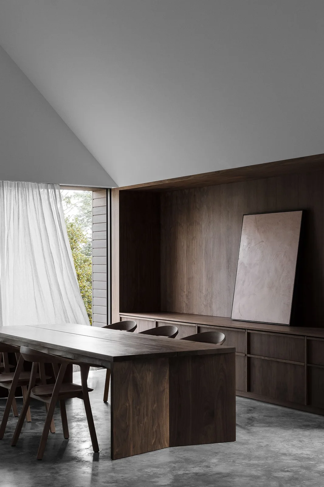 A dark wood dining table and curved wood chairs stand beside continuous wood wall panels, low matching cupboards, pale curtains and one leaning abstract artwork