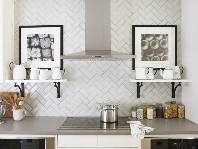 White kitchen backsplash tiles arranged in herringbone behind a metal hood, with open white shelves and dark brackets on either side.