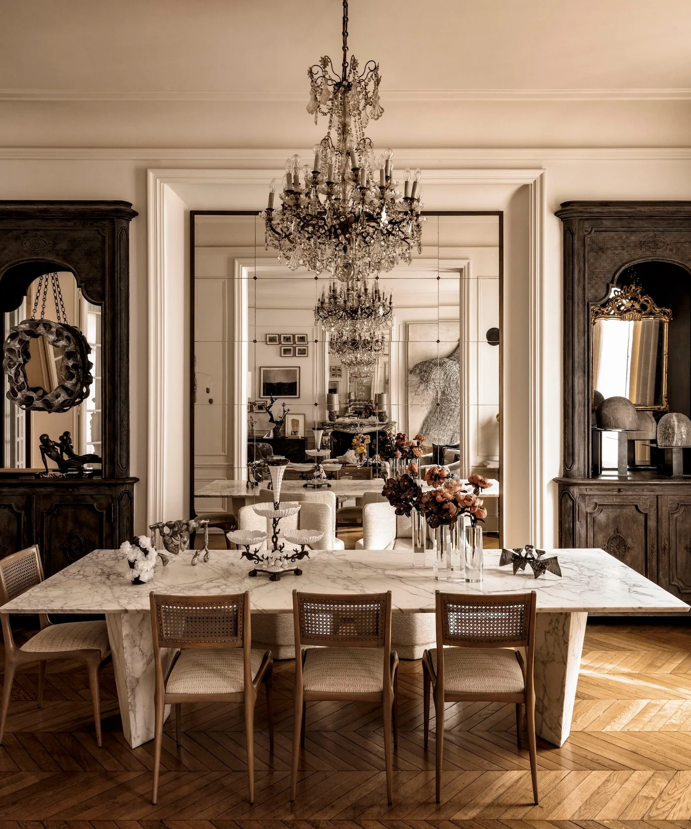 Light rectangular dining table with cane-backed chairs beside a large wall mirror, dark cabinetry and an ornate chandelier.