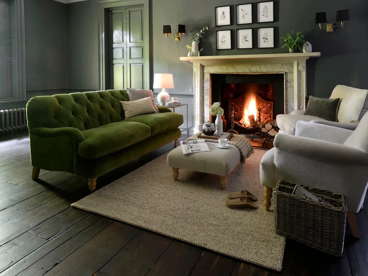 Pale traditional fireplace in a dark gray room with green sofa, light upholstered chairs and small botanical picture grid