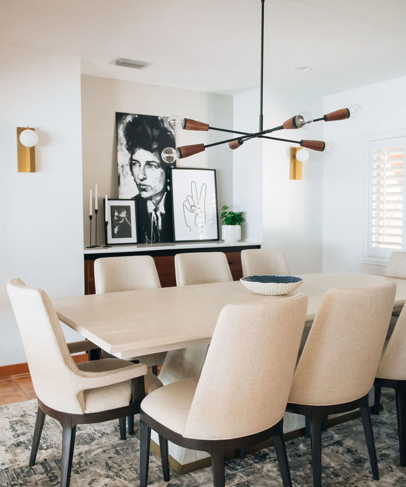 A wood-fronted sideboard with a pale upper edge stands behind a light dining table and upholstered chairs beneath leaning black-and-white artworks
