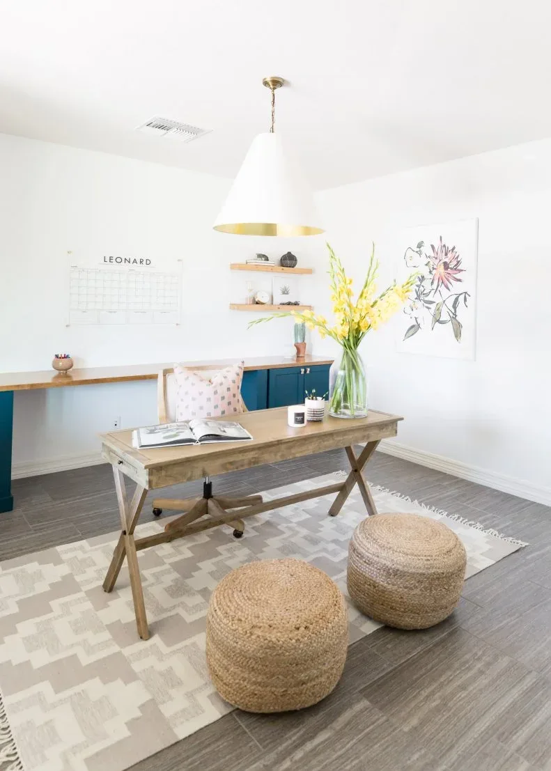 Home office with wooden trestle desk, pale patterned rolling chair, two woven poufs, geometric pale rug, blue lower cabinet and botanical wall art.