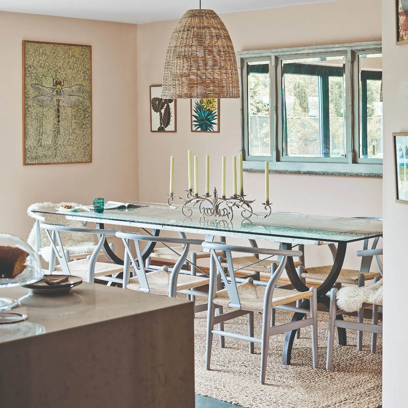 Long glass-topped dining table with pale chairs and woven seats below a large woven pendant in a peach-colored room
