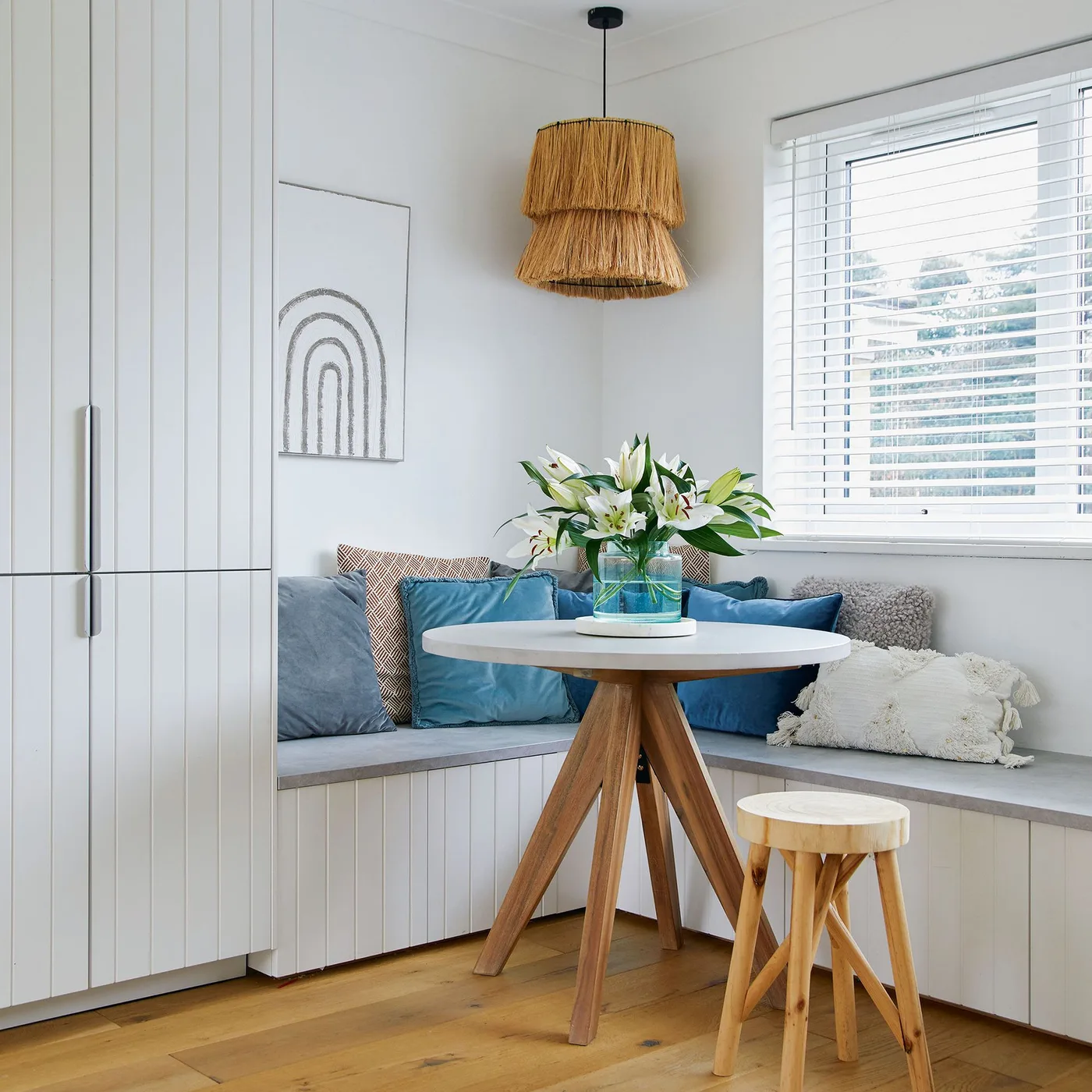 A small pale round table with wooden legs sits beside a paneled corner bench, blue cushions, a wooden stool and a textured pendant