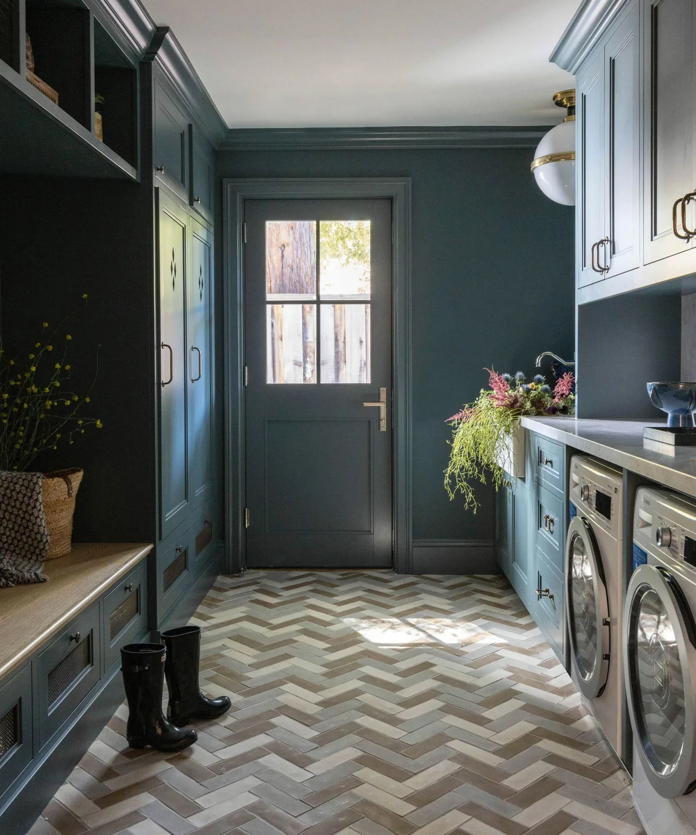 Dark blue laundry and mudroom cabinets with bench cubbies opposite side-by-side machines and pale herringbone-patterned floor