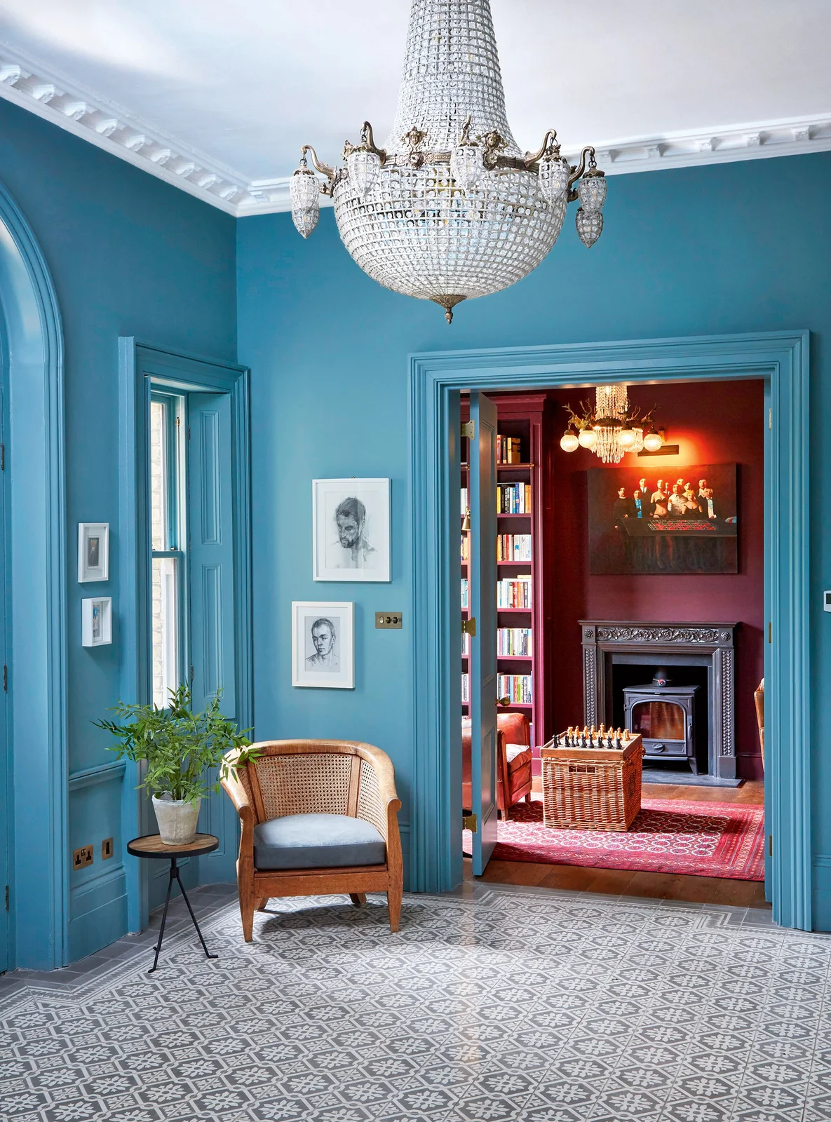 Turquoise walls and trim surround a clear decorative chandelier, framed portraits, a woven chair and a gray-and-white patterned floor