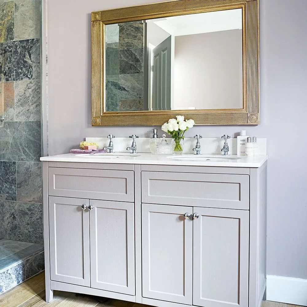 Pale paneled vanity with two basins, cabinet doors and drawers below a large gold-toned framed mirror.