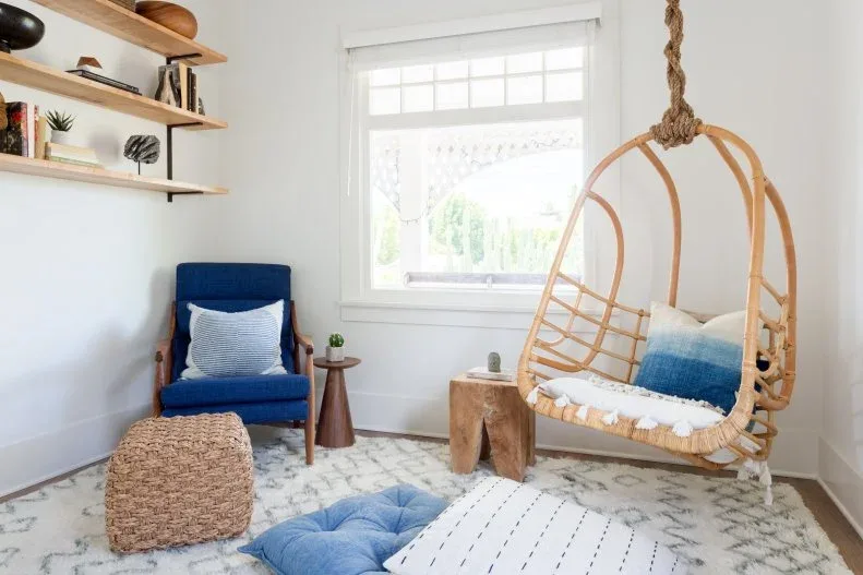 Hanging wooden chair beside a blue armchair, pale floor cushions, open shelves and a bright window.