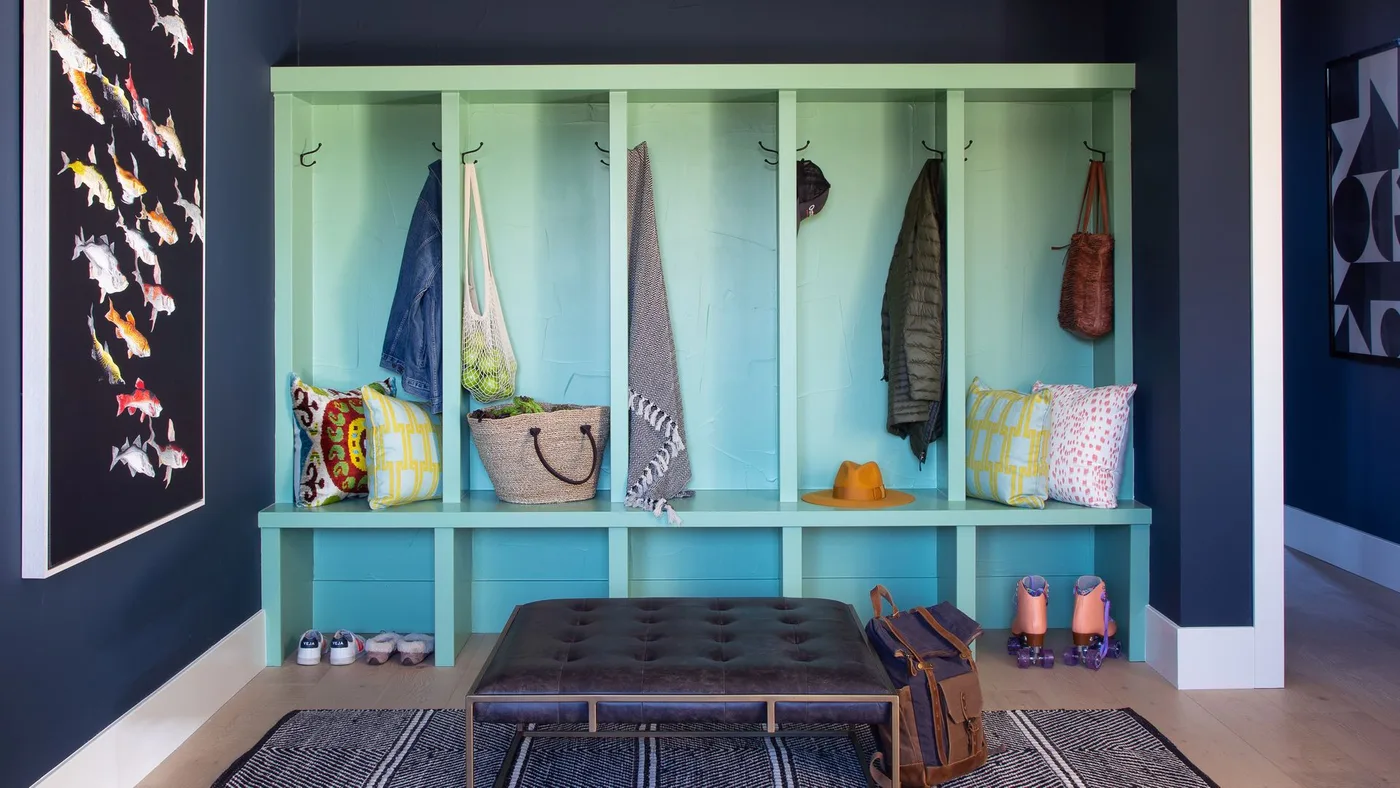 Entry mudroom with pale turquoise divided coat bays, shoe cubbies, navy walls, patterned cushions and a dark padded stool
