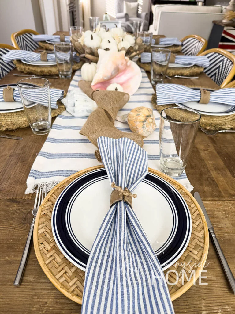A wood dining table with striped napkins holds white pumpkins, starfish shapes and a pink conch shell on a small raised stand