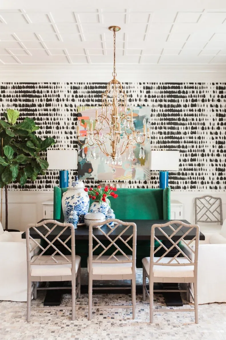 Gold-toned curving chandelier with clear hanging drops above a dark table, green bench, pale lattice-back chairs, and black patterned wallpaper.