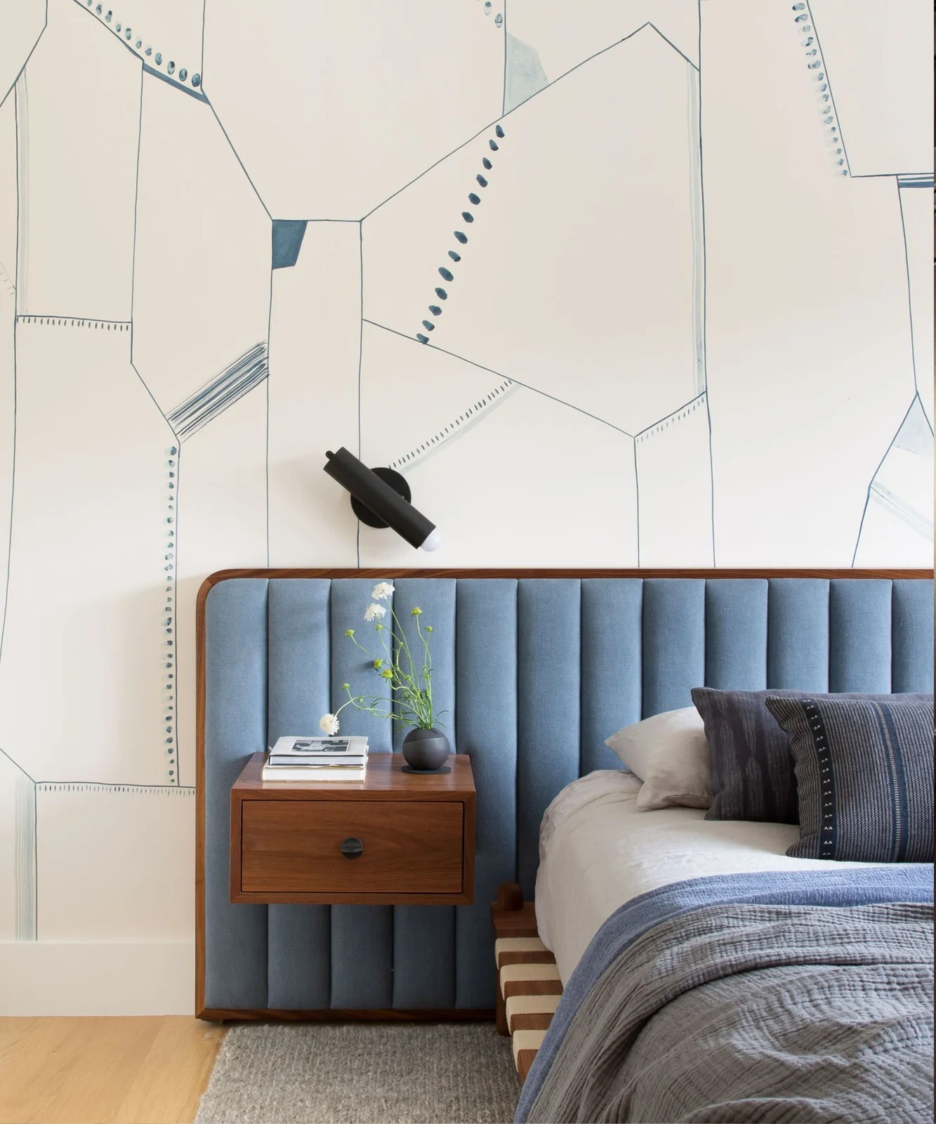 A blue paneled upholstered headboard stands against pale geometric wallpaper beside a floating wooden drawer, dark wall light and blue bed layers