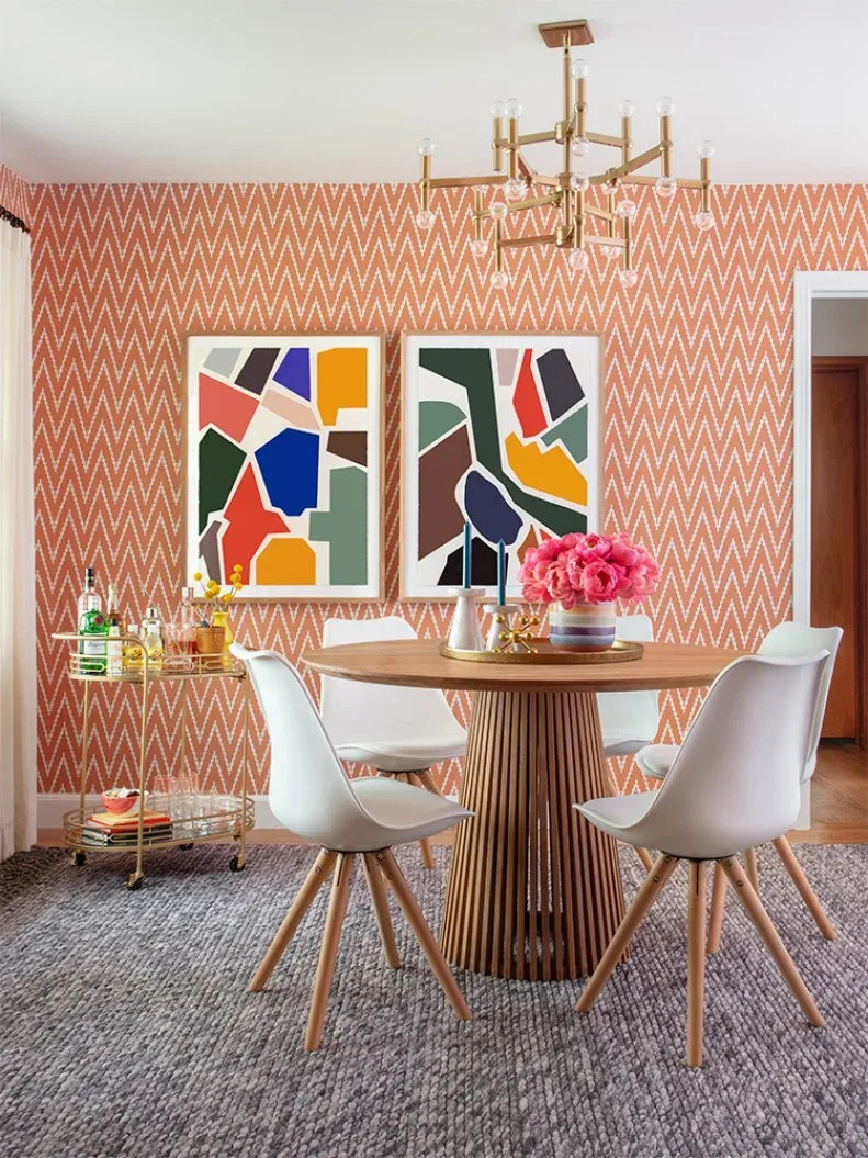 Coral-and-white chevron dining wall with two colorful abstract pictures, round wood table, pale chairs, gold-tone chandelier, and a small drinks cart.