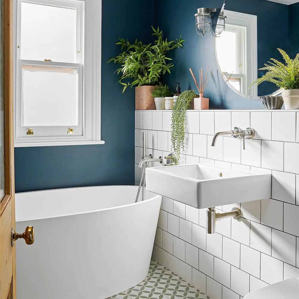 An inky blue wall surrounds a white window above white rectangular tiles beside a bath and wall-mounted sink
