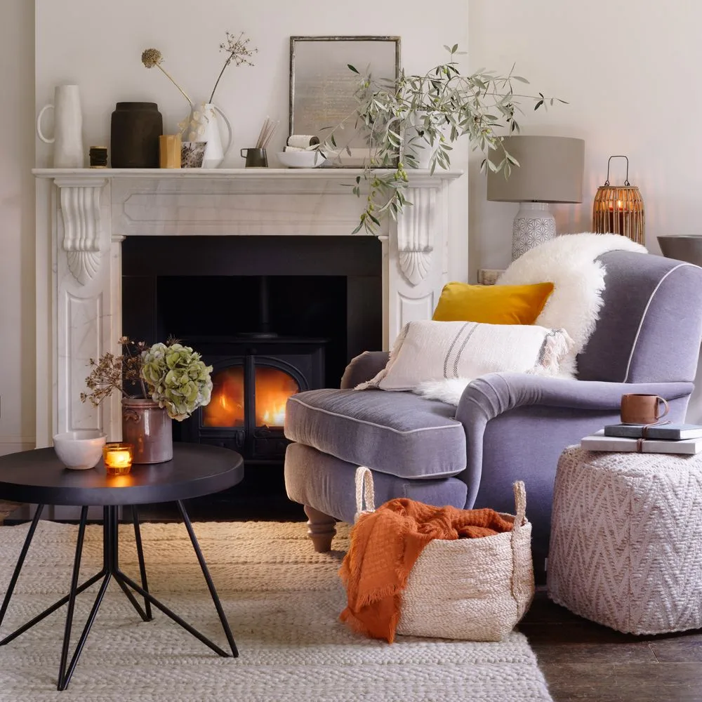 Pale fireplace with gray upholstered armchair, woven blanket basket and round black coffee table