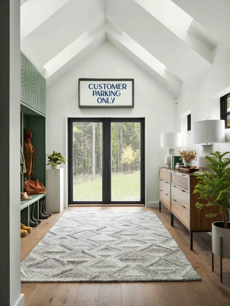 Light high-ceiling mudroom with green coat storage, wooden drawer cabinet, plant and large pale geometric rug.