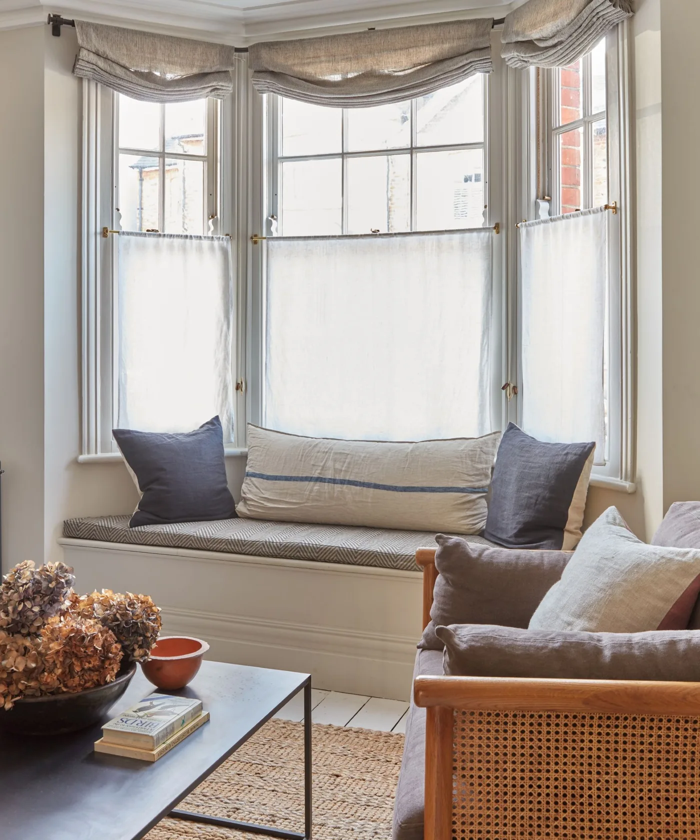 A pale bay-window seat with simple cushions stands beside a woven-backed wood chair, a dark coffee table and light window curtains