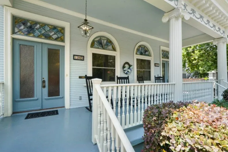 Pale porch with blue double door, dark rocking chairs, white railing and planted bed beside the steps