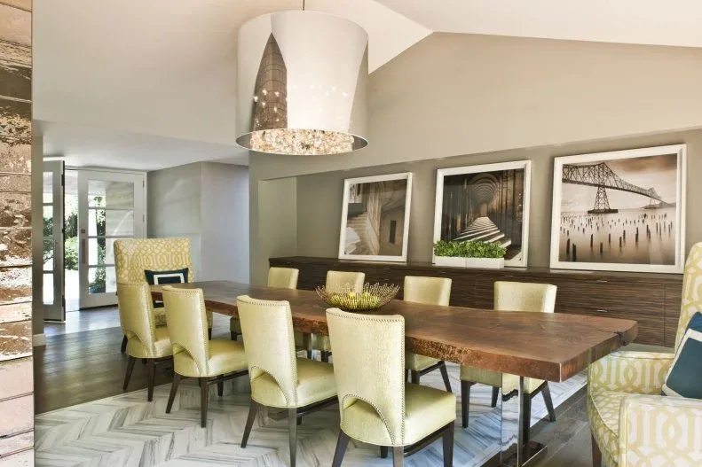 Pale green upholstered dining chairs with dark legs and curved back openings beside a long wooden table, with framed photographs and a shaded pendant.