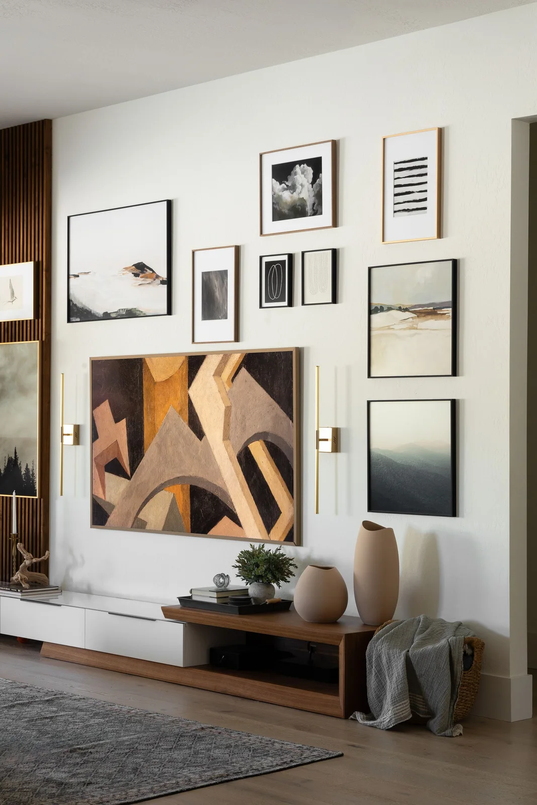 An art-displaying television sits within a gallery of framed pictures above a low cabinet between slender gold-toned wall lights
