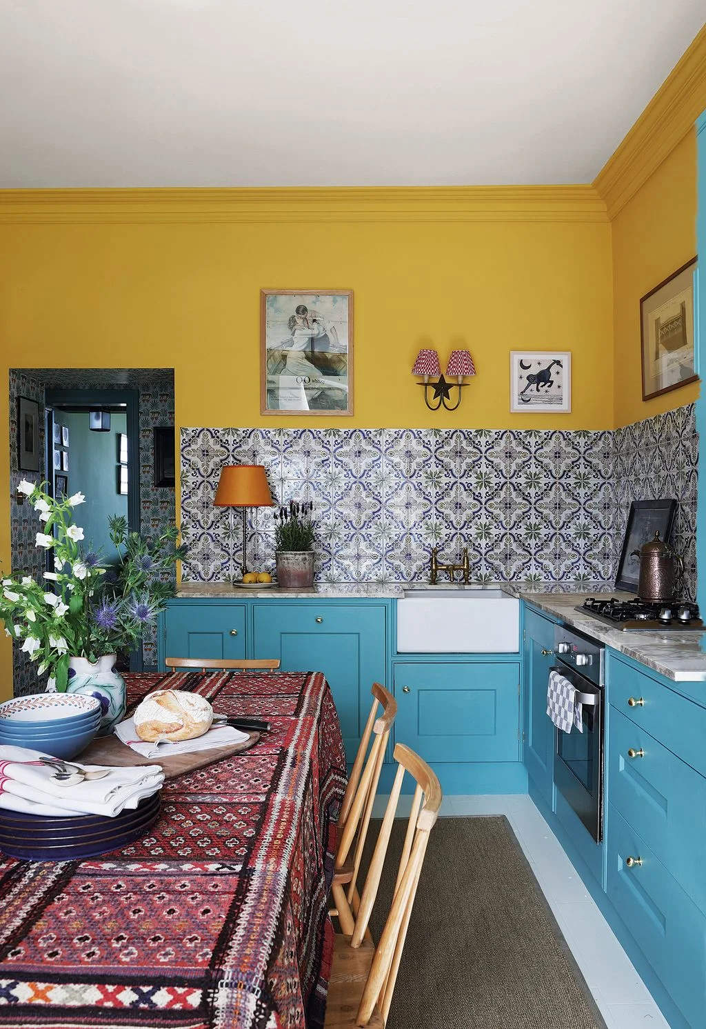 Yellow kitchen walls sit above blue-and-white patterned tiles and turquoise cabinets beside a red patterned tablecloth and framed artworks