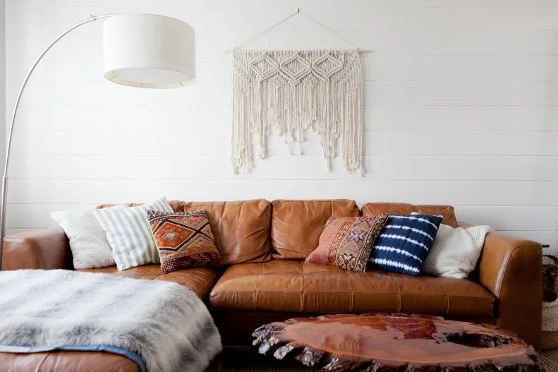 Brown sofa with patterned cushions, pale throw, macrame wall hanging and a curved floor lamp.