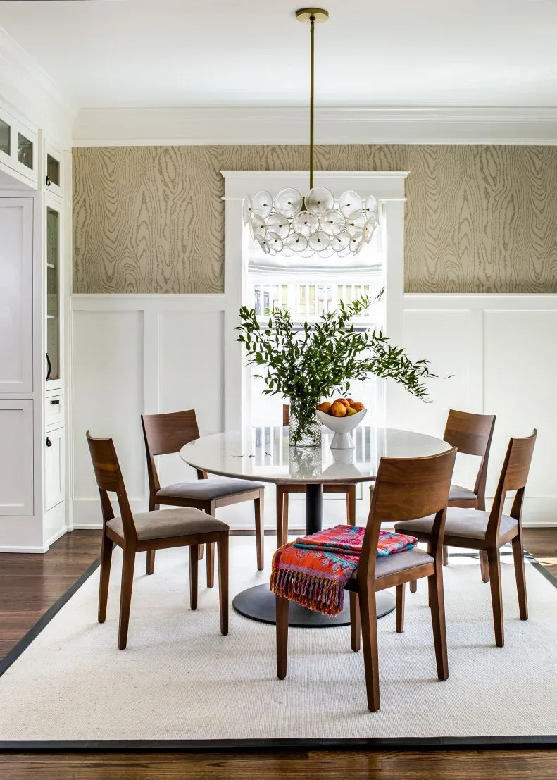 Rounded chandelier with clear circular elements and a gold-toned stem above a round pale table, wooden chairs, patterned wall covering, and white paneling.