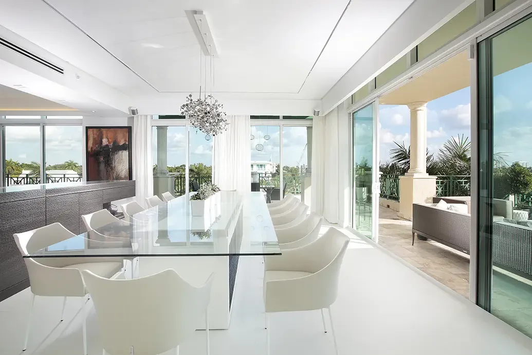 A long clear glass dining table on a pale central support stands between white curved chairs beside broad sliding doors and dark lower storage