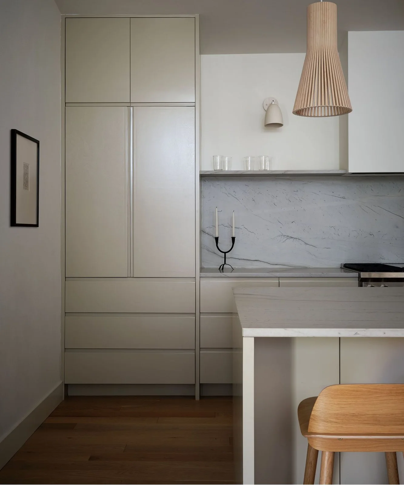 Pale flat-front cabinets surround a lightly veined backsplash beside a wooden stool, narrow dark shelf and textured tapered pendant