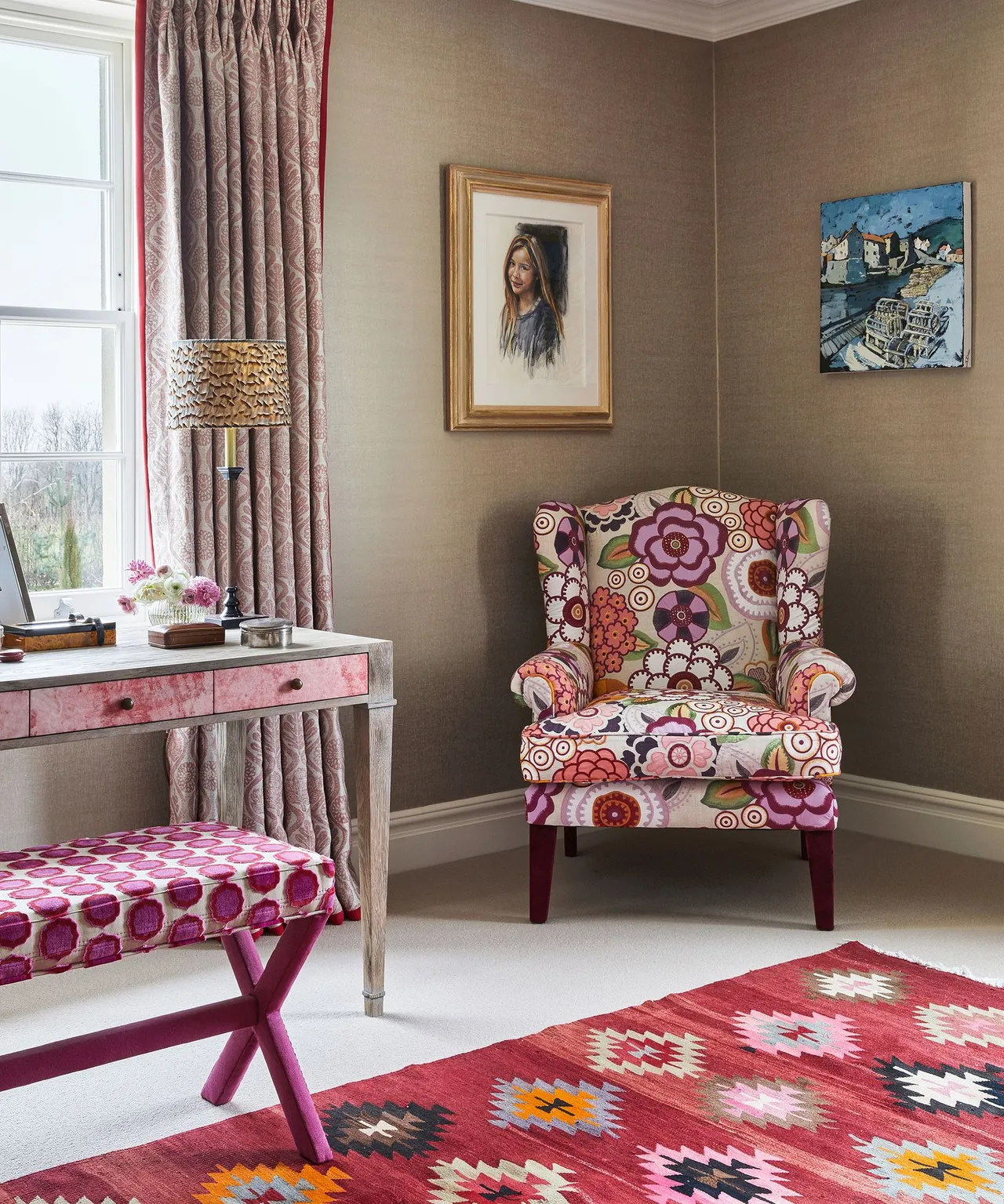 Bedroom sitting area with taupe walls, patterned floral armchair, pink drawer dressing table, upholstered folding stool, framed pictures and red patterned rug.