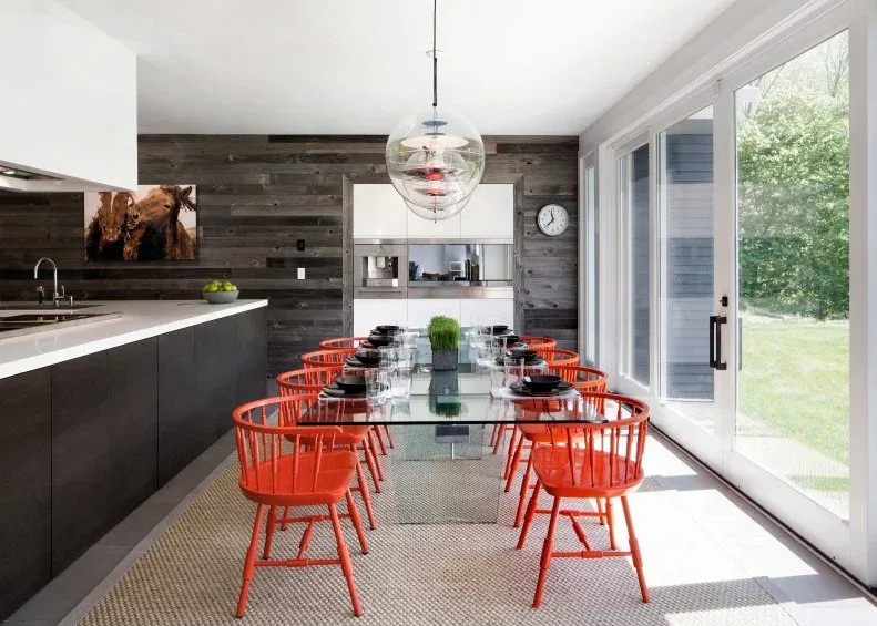Open kitchen dining area with a gray-brown horizontal wood feature wall, glass table, orange chairs, glass pendants, pale ceiling, and large sliding doors.